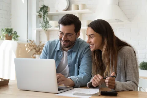 Smiling man and woman sit together at a table, looking at a laptop screen with notebooks and papers in front of them in a bright, cozy kitchen.
