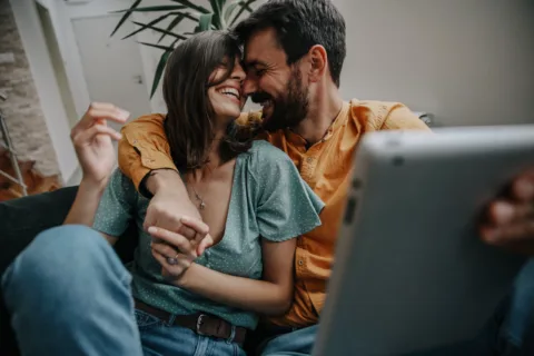 A happy couple sits close together on a couch, laughing and holding hands, with one holding a tablet and a plant visible in the background.