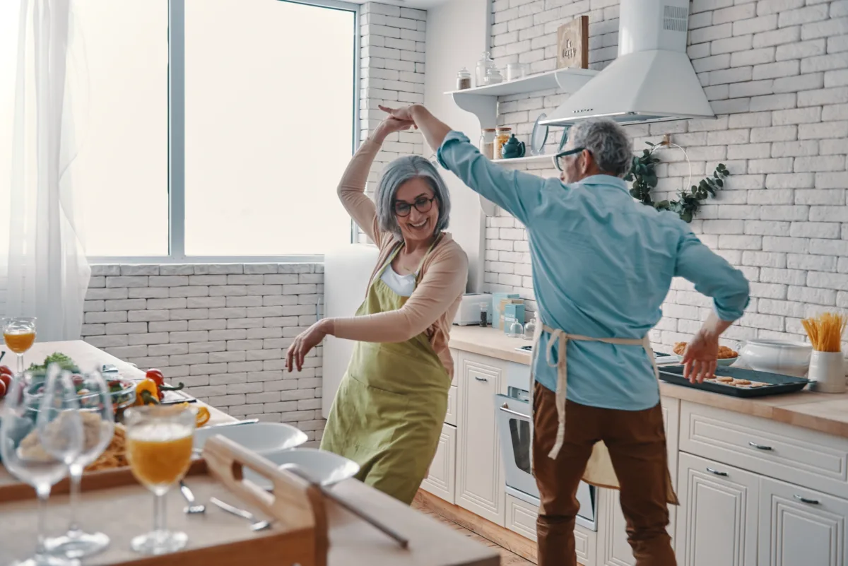 An older couple wearing aprons dance and smile together in a bright, modern kitchen with food and drinks on the counter.