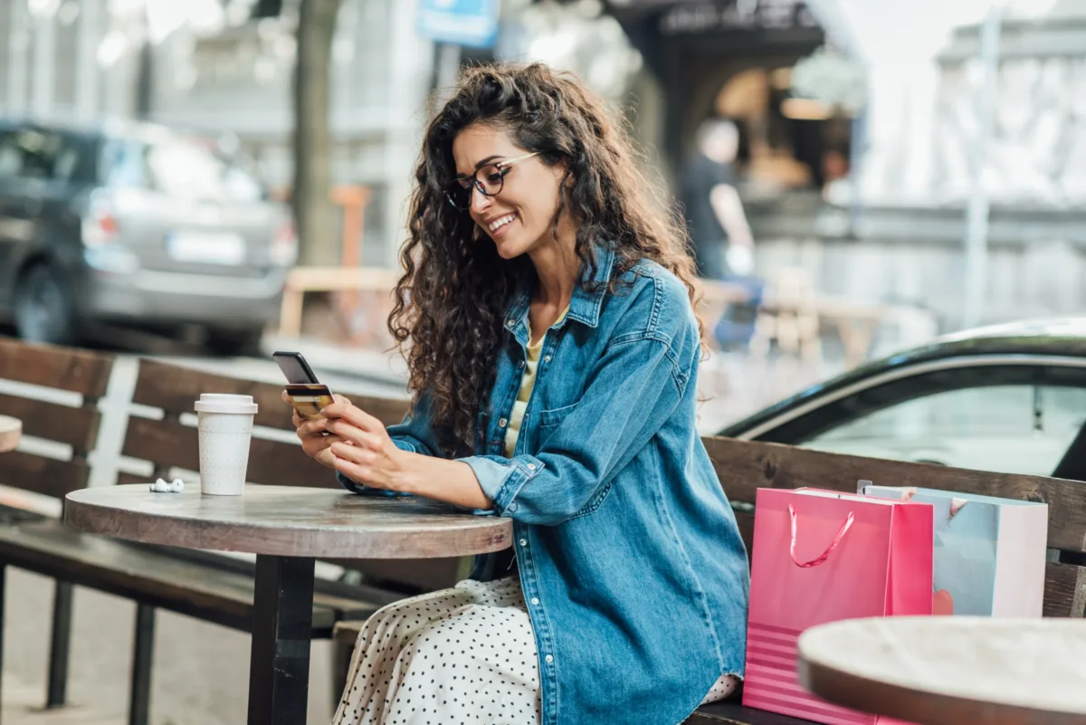 A woman with curly hair and glasses sits at an outdoor café table, smiling at her phone. She wears a denim jacket, and there are shopping bags and a coffee cup on the table.