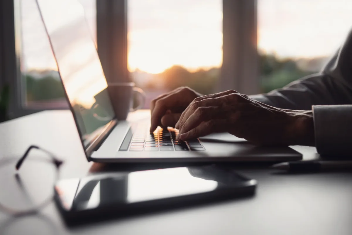 Close-up of hands typing on a laptop keyboard, with a tablet, eyeglasses, and a mug on a desk, and a window showing a sunset in the background.