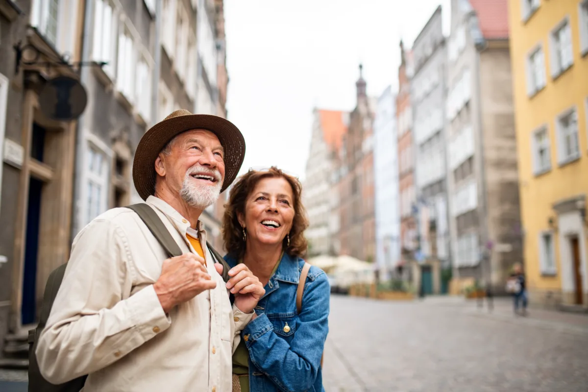 An older couple smiles and looks up while exploring a cobblestone street in a historic European city, surrounded by colorful old buildings.