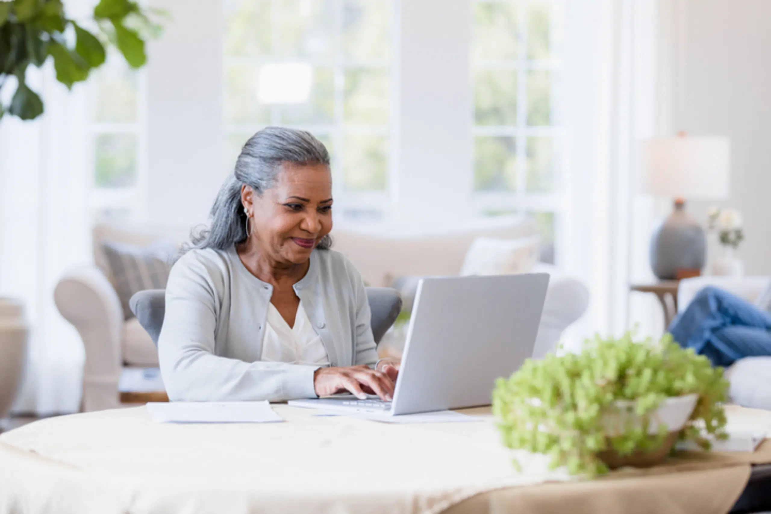 An older woman with gray hair smiles while using a laptop at a table in a bright, cozy living room with plants and natural light.