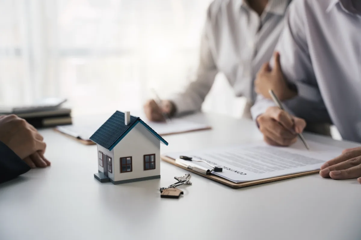 Three people sit at a table, signing documents with a clipboard, house model, and keys in focus, suggesting a real estate or home-buying transaction.