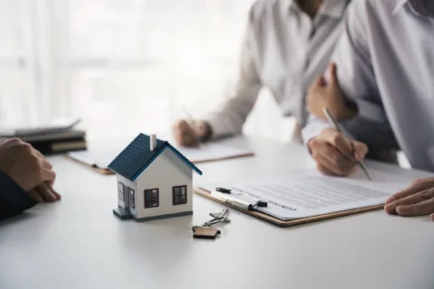 Three people sit at a table, signing documents with a clipboard, house model, and keys in focus, suggesting a real estate or home-buying transaction.
