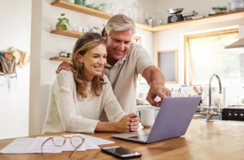 Smiling older couple in a kitchen, looking at a laptop together; the man is pointing at the screen while the woman sits beside him. Papers and a smartphone are on the counter.