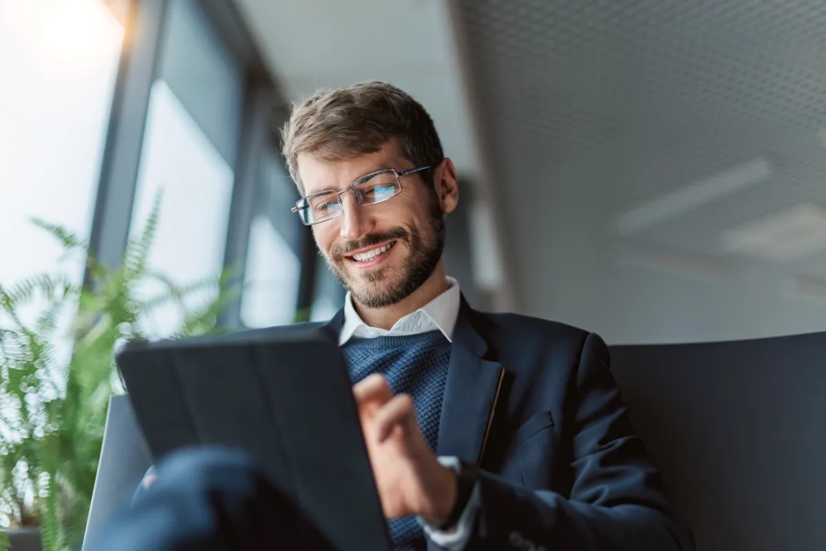 A smiling man in a suit and glasses uses a tablet while sitting indoors near large windows with natural daylight.