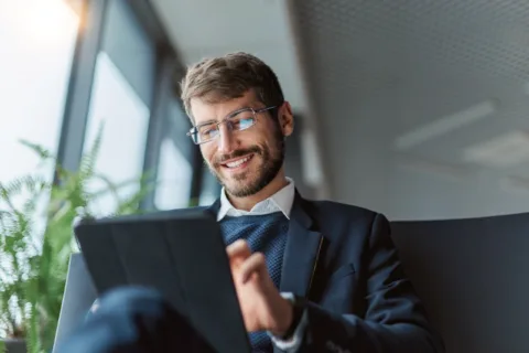 A smiling man in a suit and glasses uses a tablet while sitting indoors near large windows with natural daylight.