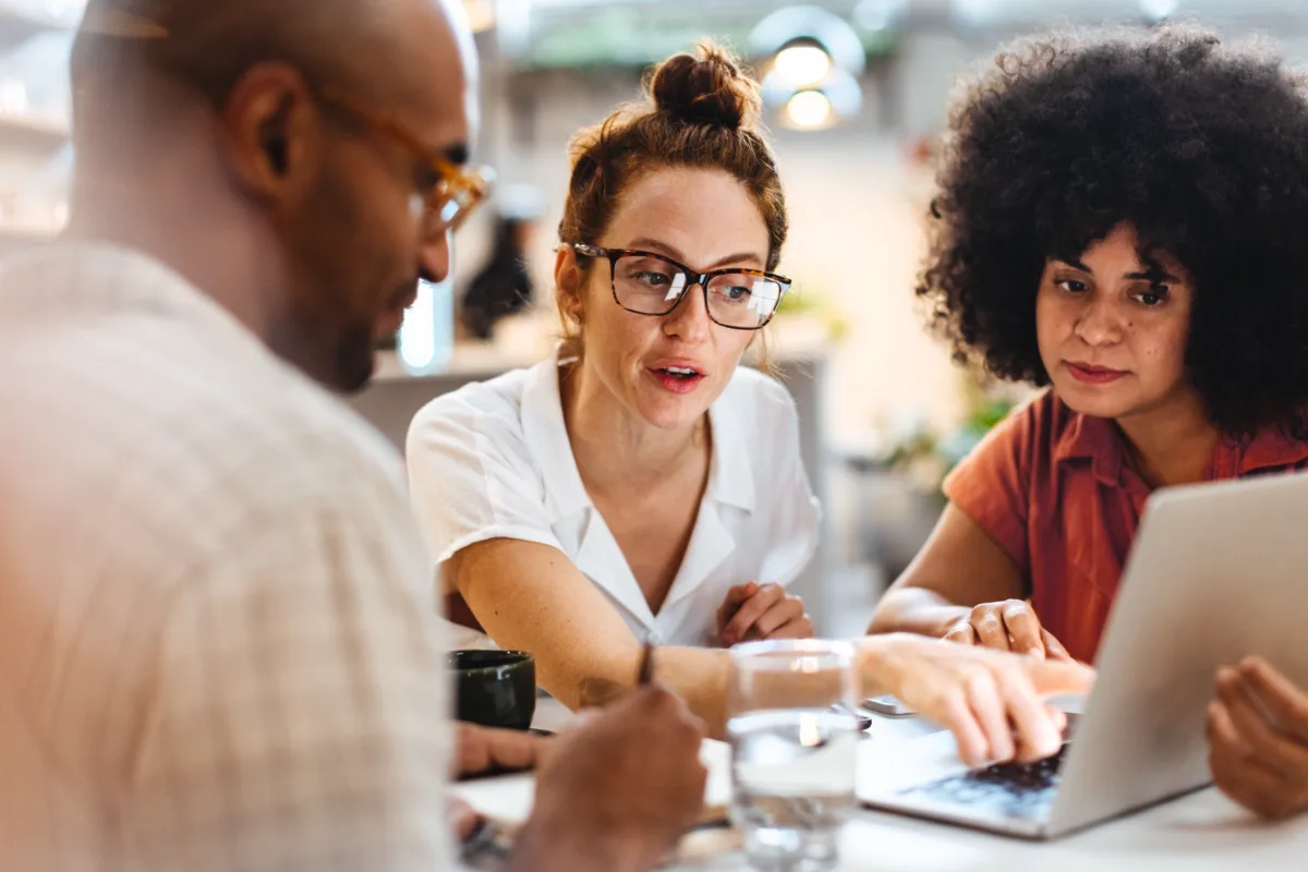 Three people sit at a table, collaborating and looking at a laptop screen. One person is pointing at the screen while the others listen and watch attentively.