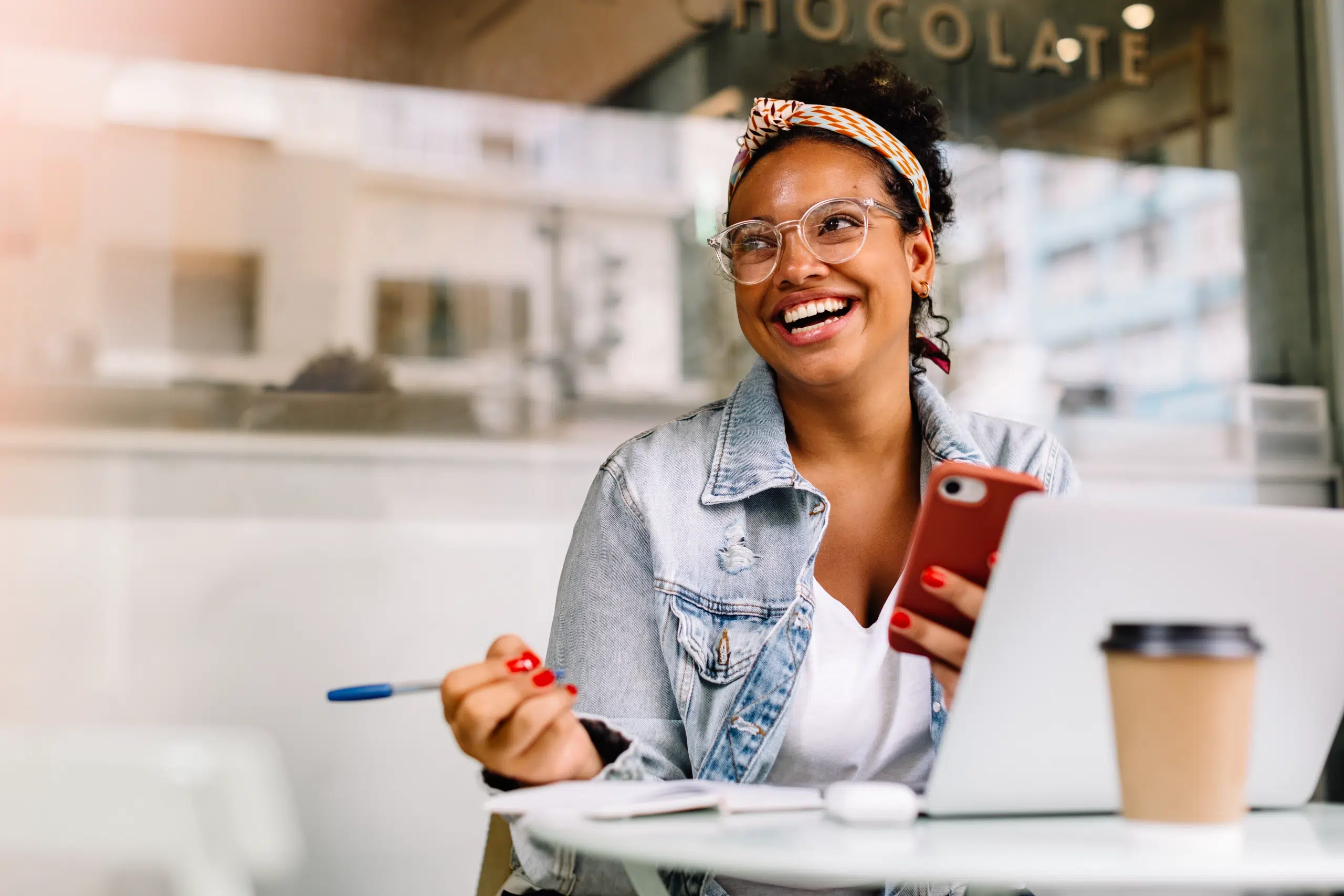 Smiling woman wearing glasses holds a phone and pen, seated at a table outdoors with a laptop and coffee cup.