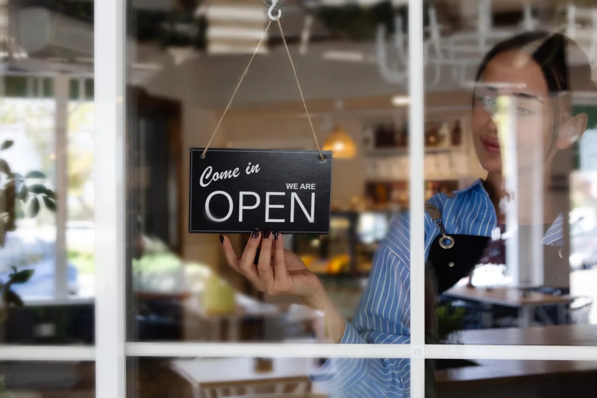 A woman in an apron hangs an Open, Come in sign on the glass door of a well-lit café or shop, inviting customers inside.