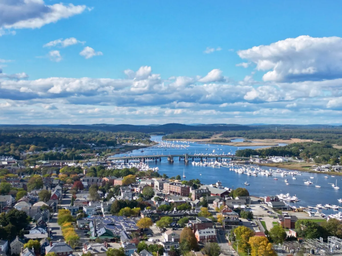 Aerial view of a riverside town with boats docked along the water, a bridge crossing the river, and a mix of trees and buildings under a partly cloudy sky.