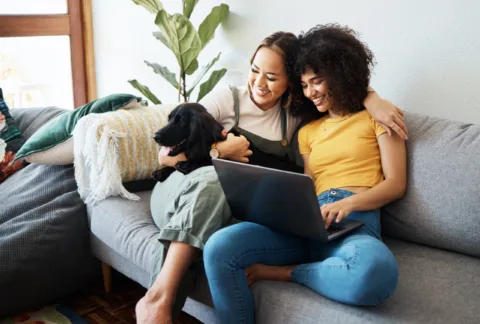 Two women sit on a couch with a black dog, smiling and looking at a laptop together in a cozy living room.