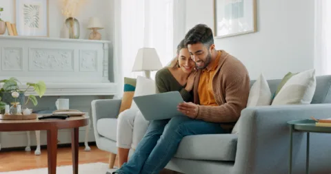A smiling couple sits close together on a couch, looking at a laptop. The living room is bright and decorated with a piano, lamp, and framed art.