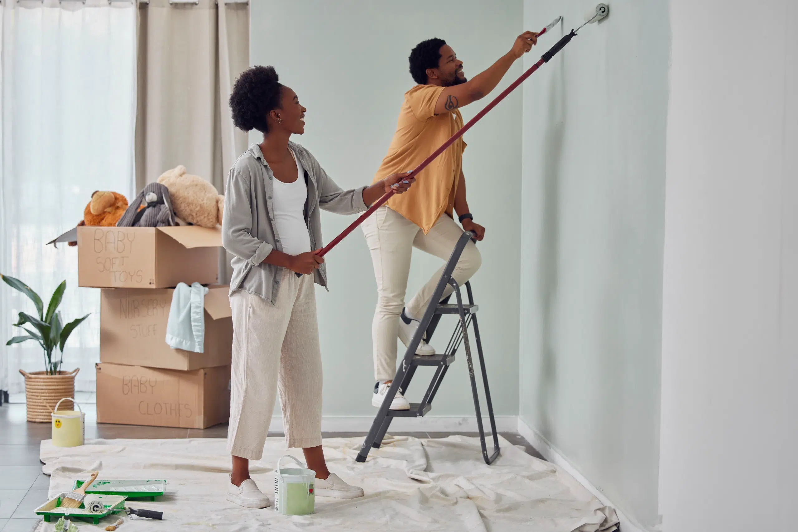 A couple paints a light-colored wall together; one stands on a stepladder with a roller while the other holds the extension pole. Cardboard boxes and a plant are in the background.