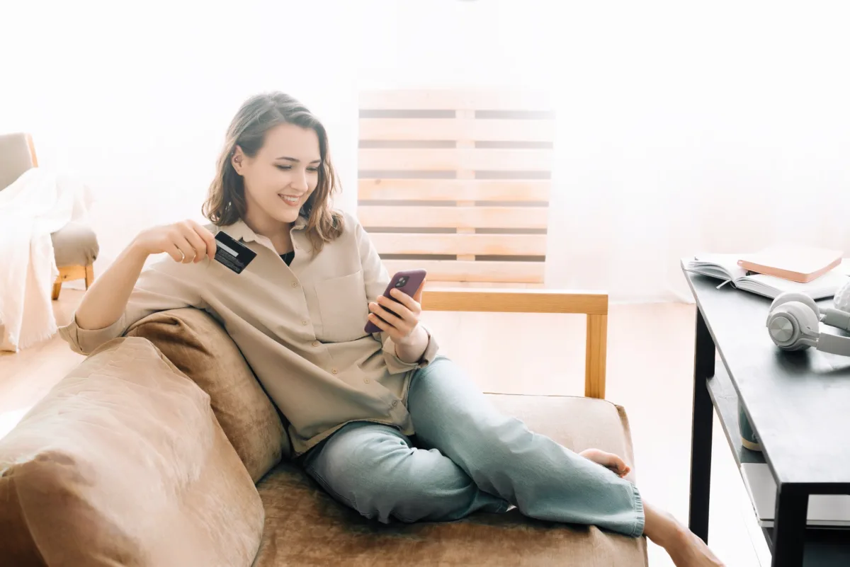 A woman sits on a couch, smiling while holding a credit card and using her smartphone. A table with headphones, a book, and a notebook is nearby.