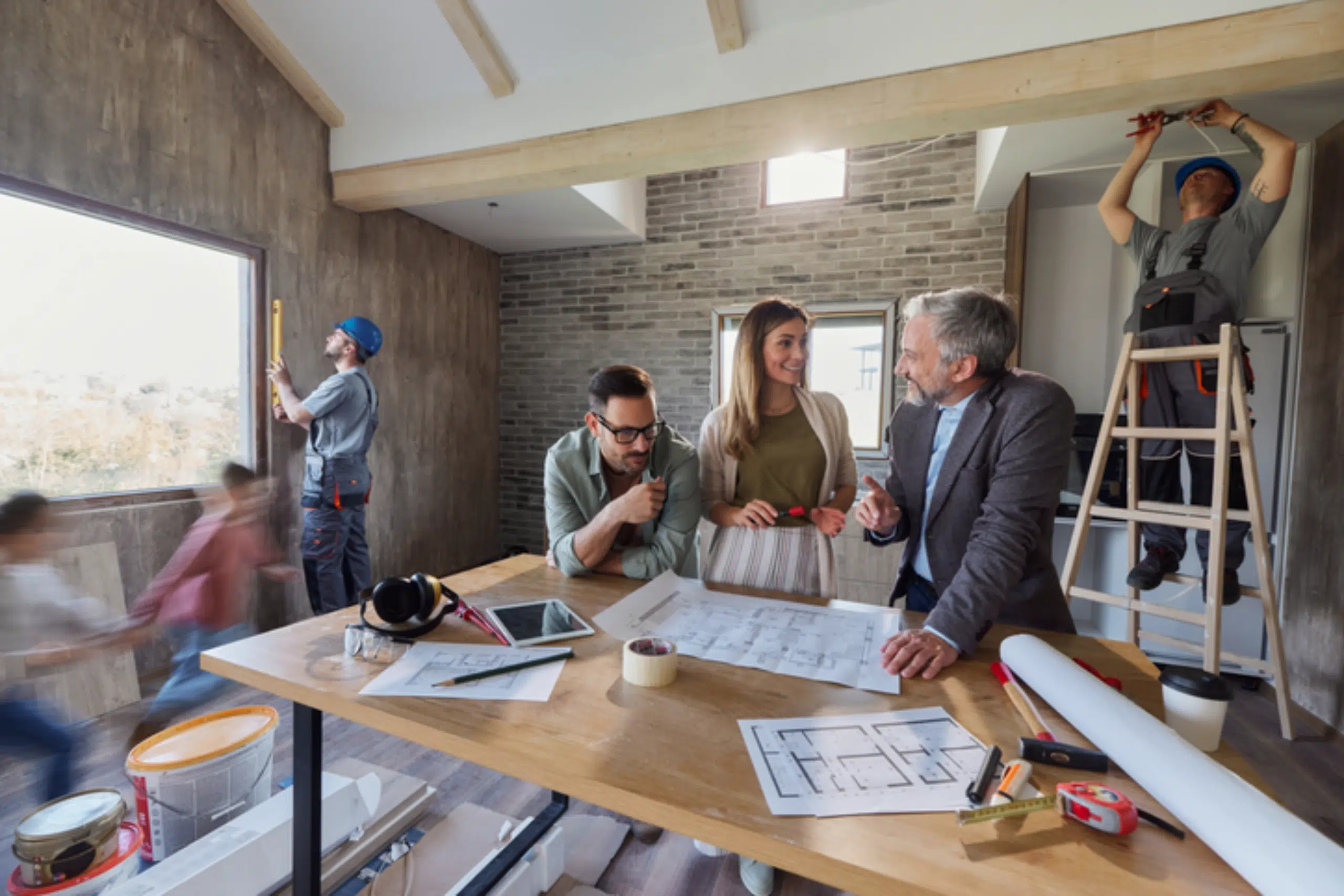 Four people discuss blueprints at a table in a modern room under renovation, while two workers measure and work on the walls and window in the background.