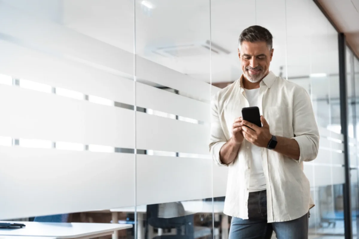 A smiling man in a light shirt stands in a modern office, looking at his smartphone near a glass wall with frosted horizontal stripes.