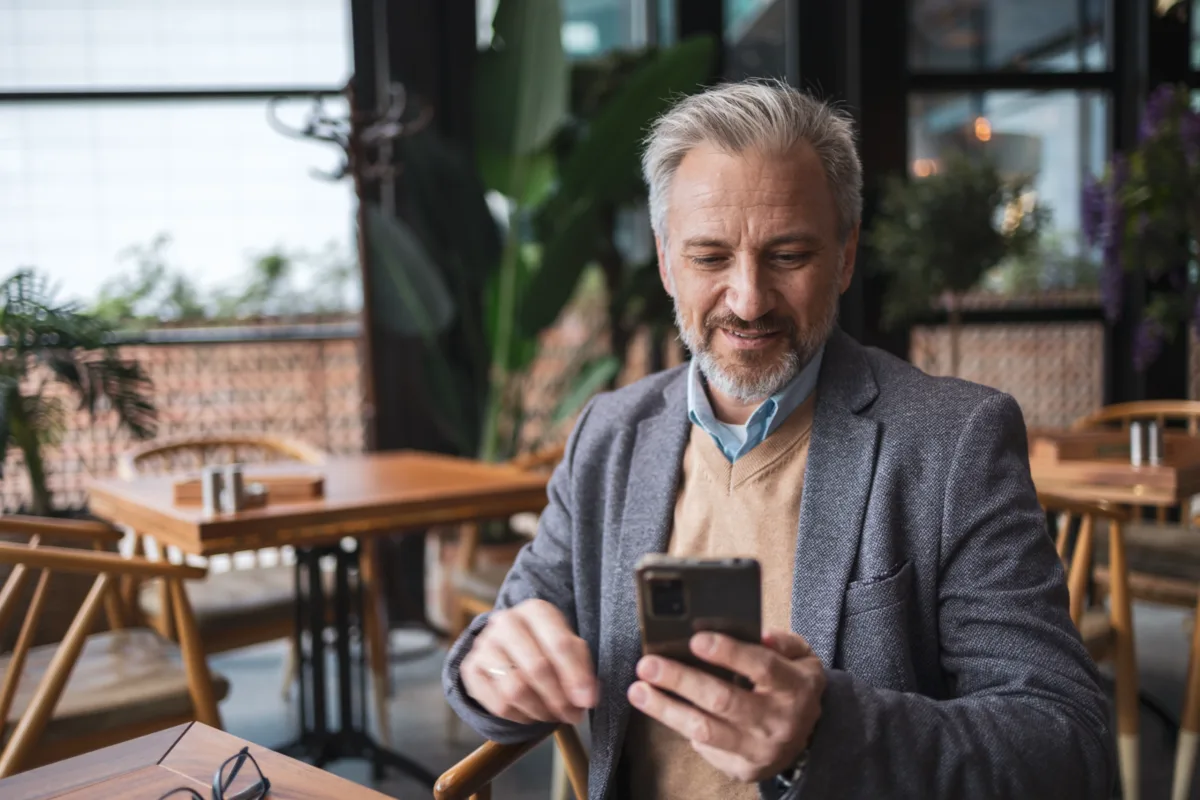 A smiling man with gray hair and a beard sits in a cafe, wearing a blazer and sweater, looking at his smartphone.
