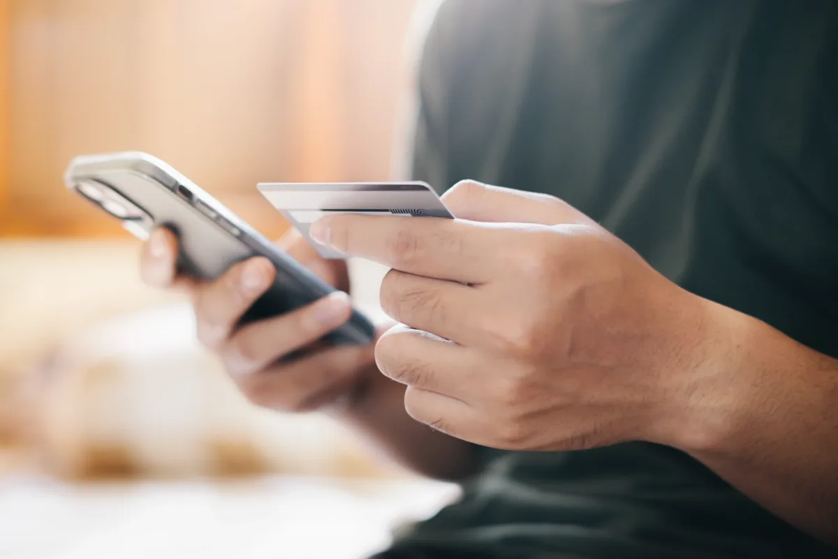 A person holding a smartphone in one hand and a credit card in the other, appearing to make an online payment or purchase.