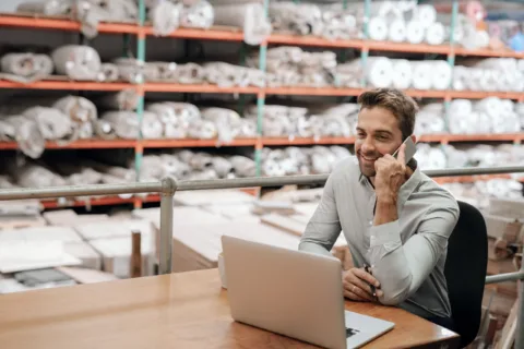 Man sitting at a desk in a warehouse, smiling while talking on the phone, with an open laptop in front of him and shelves of rolled materials in the background.