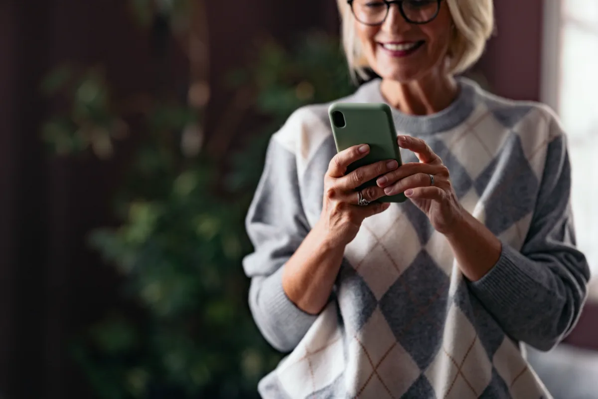 Smiling woman wearing glasses and an argyle sweater uses a smartphone indoors, with a blurred plant in the background.