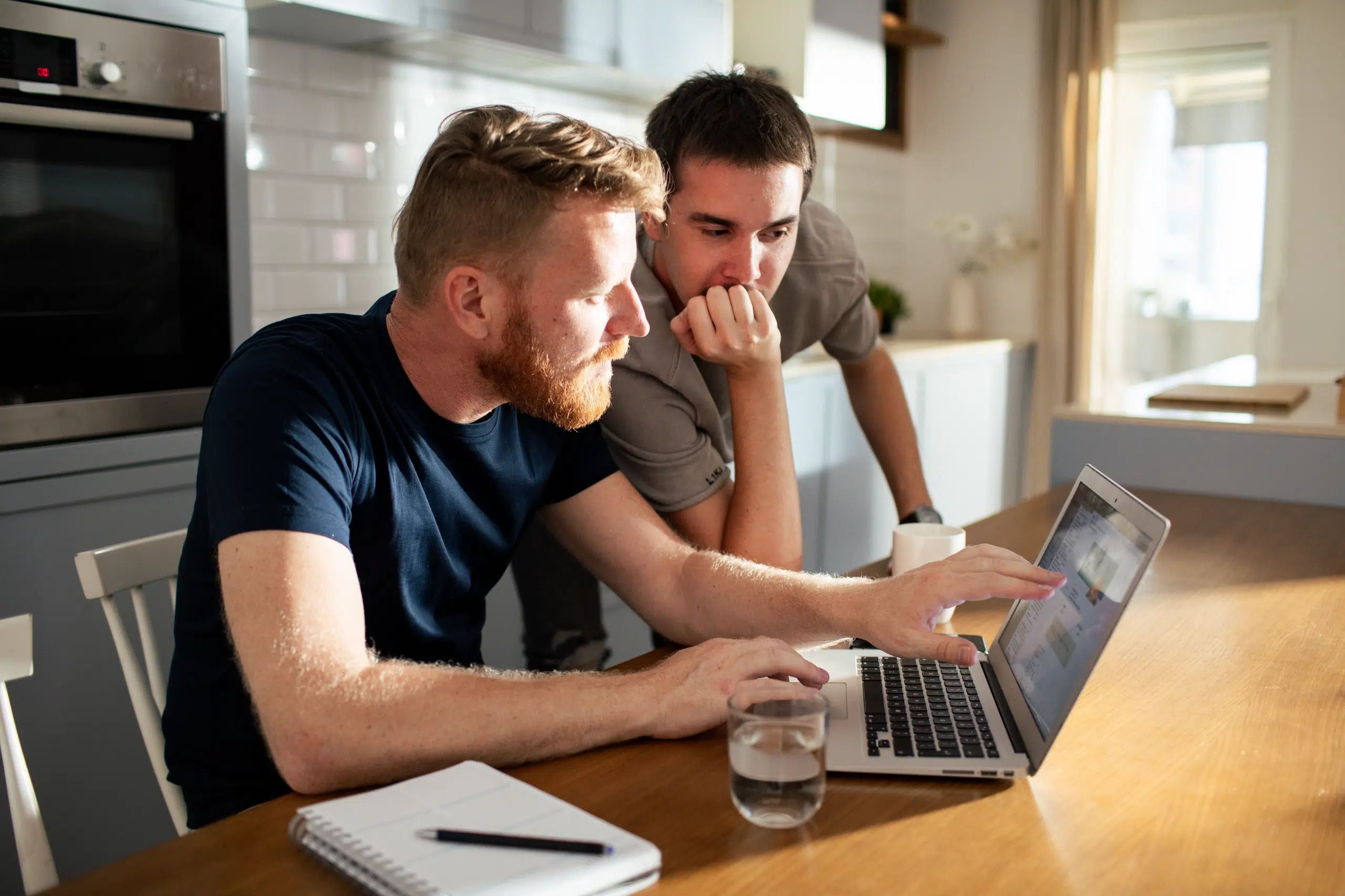 Two men sit at a kitchen table, looking intently at a laptop screen. One points at the screen while the other leans in. A notebook, pen, and glass of water are on the table.