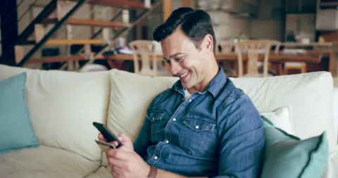 A man in a denim shirt sits on a couch, smiling while looking at his smartphone in a cozy, sunlit living room.