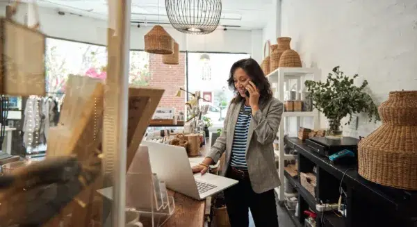 A woman stands in a boutique shop, talking on the phone while working on a laptop at the counter, surrounded by woven baskets and decorative items.