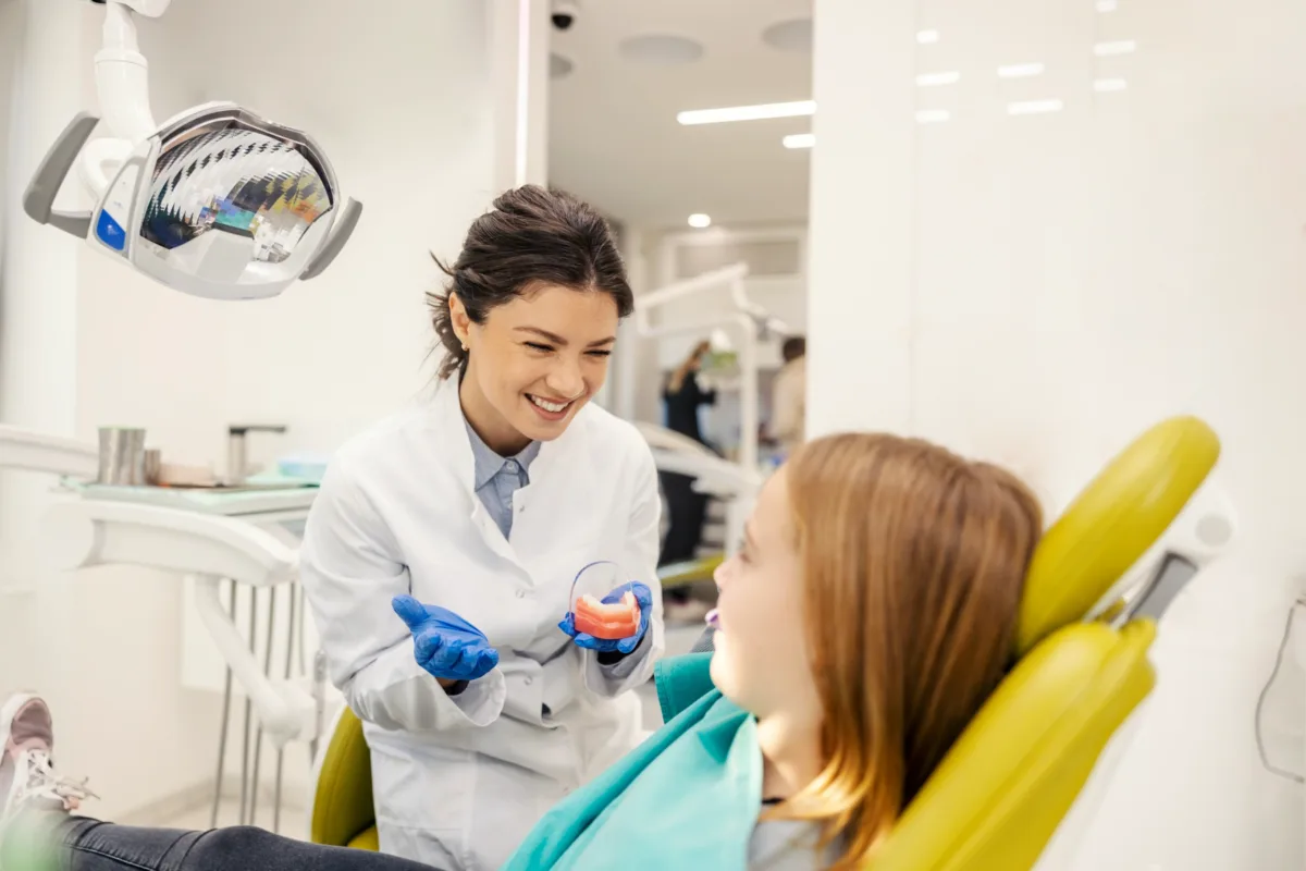 A smiling dentist wearing gloves shows a dental model to a young patient sitting in a dental chair in a bright clinic.