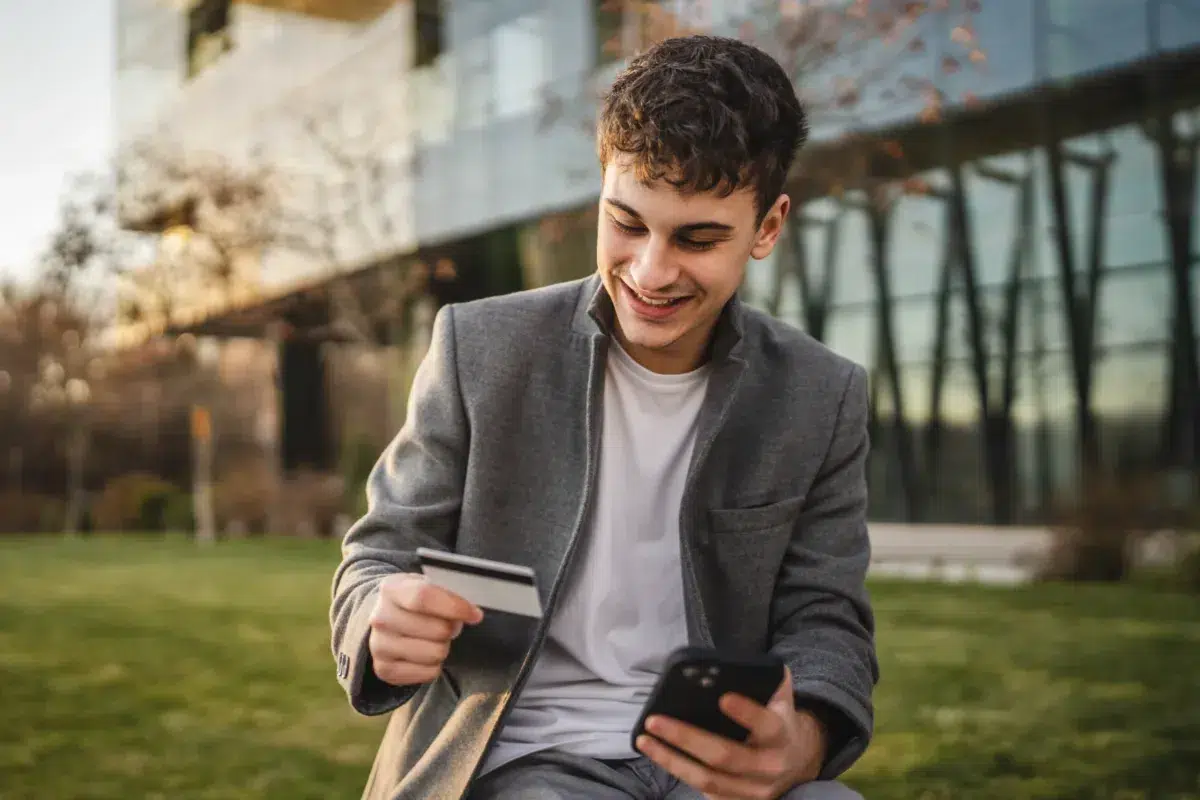 A young man sits outside, smiling while holding a credit card in one hand and a smartphone in the other. He is dressed in a gray jacket and white shirt.