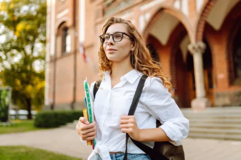 A young woman with glasses and curly hair holds books and a backpack, standing outside a brick university building, looking confident.