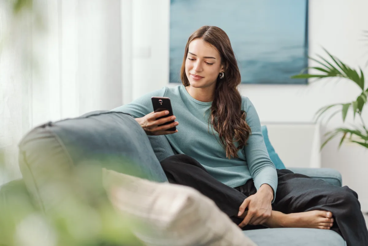 A young woman sits barefoot on a couch, smiling as she looks at her smartphone, with a plant and a painting in the background.