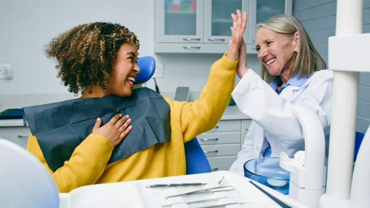 A patient and a dentist share a joyful high-five in a dental office, both smiling warmly. Dental tools and equipment are visible in the foreground.