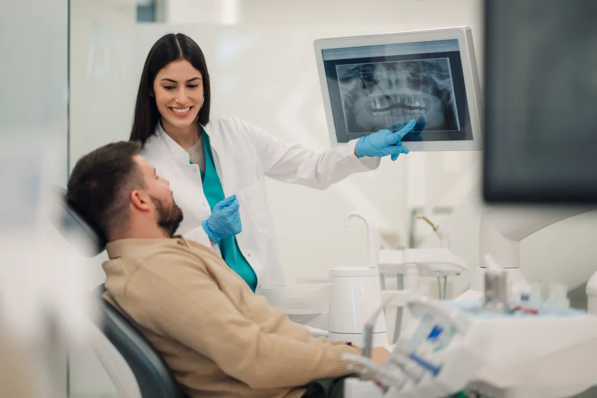 A female dentist shows a dental X-ray on a screen to a male patient sitting in a dental chair, both smiling in a modern dental clinic.