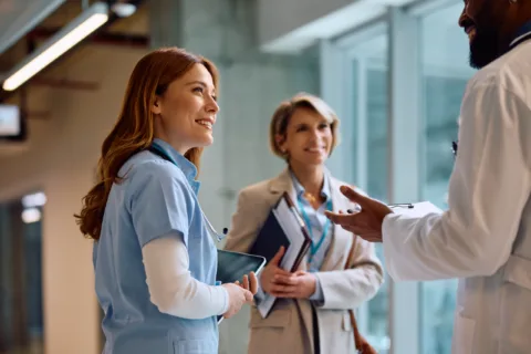 Three healthcare professionals stand indoors, smiling and talking; one wears light blue scrubs, another wears a blazer, and the third is in a white coat holding a clipboard.