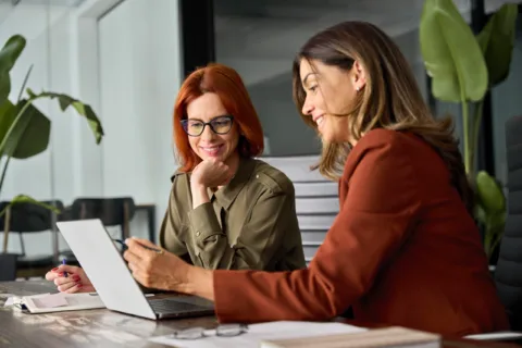 Two women sit at a desk in an office, smiling and looking at a laptop screen while discussing work. Papers and a notebook are on the table, and large green plants are in the background.