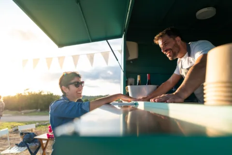 A smiling woman in sunglasses chats with a cheerful man working at an outdoor food truck during sunset. Bunting hangs above them, and drinks are visible on the counter.