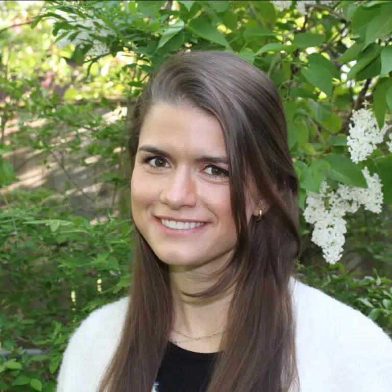 A woman with long brown hair, wearing a white sweater, smiles in front of green leafy bushes and white lilac flowers.