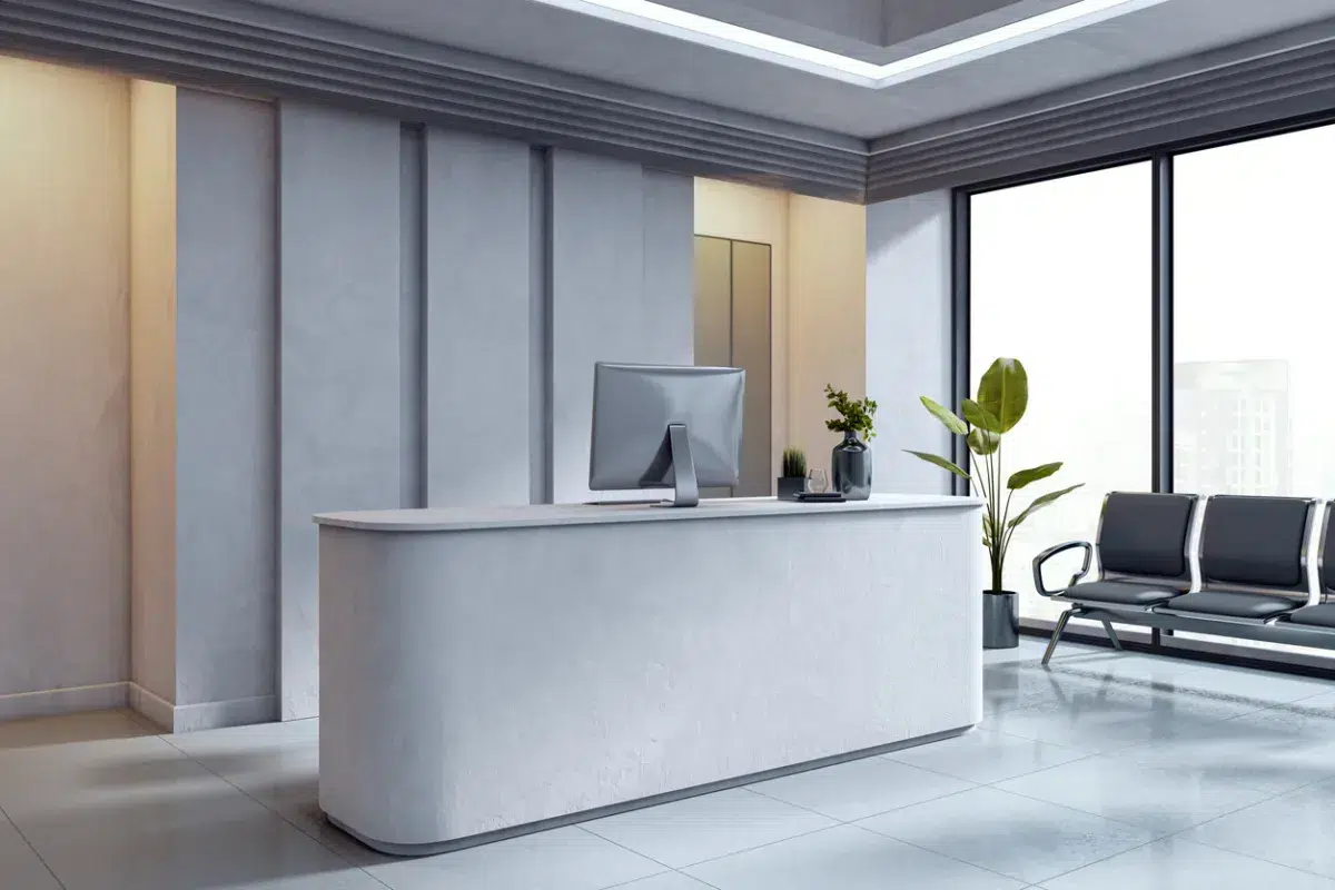A modern, minimalist office reception area with a curved desk, computer, potted plants, and large windows letting in natural light. Black chairs line the wall near the windows.
