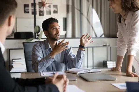 Three colleagues discuss commercial lending in Massachusetts at an office table—one man speaks animatedly, a woman stands and listens, while another sits with hands folded.