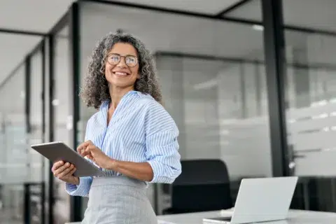 Smiling woman with curly gray hair and glasses holds a tablet in a modern office, standing by a desk with an open laptop, ready for a treasury management meeting.