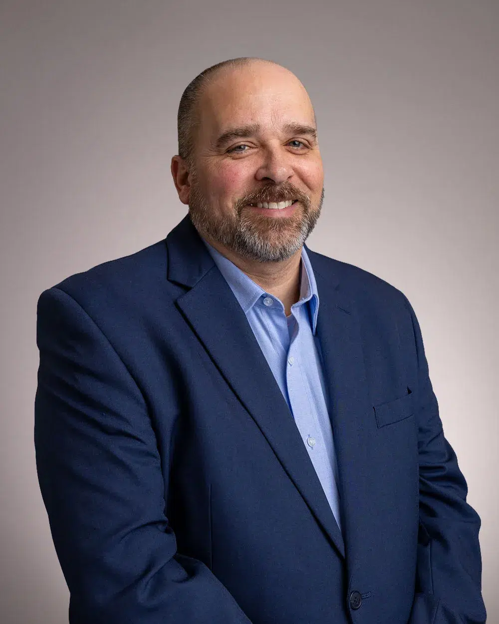 A man with a beard and short hair, wearing a blue suit jacket and light blue shirt, smiles while posing in front of a neutral background.
