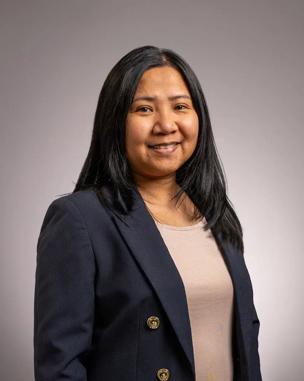 A woman with long black hair, wearing a navy blazer and beige top, smiles at the camera against a plain gray background.