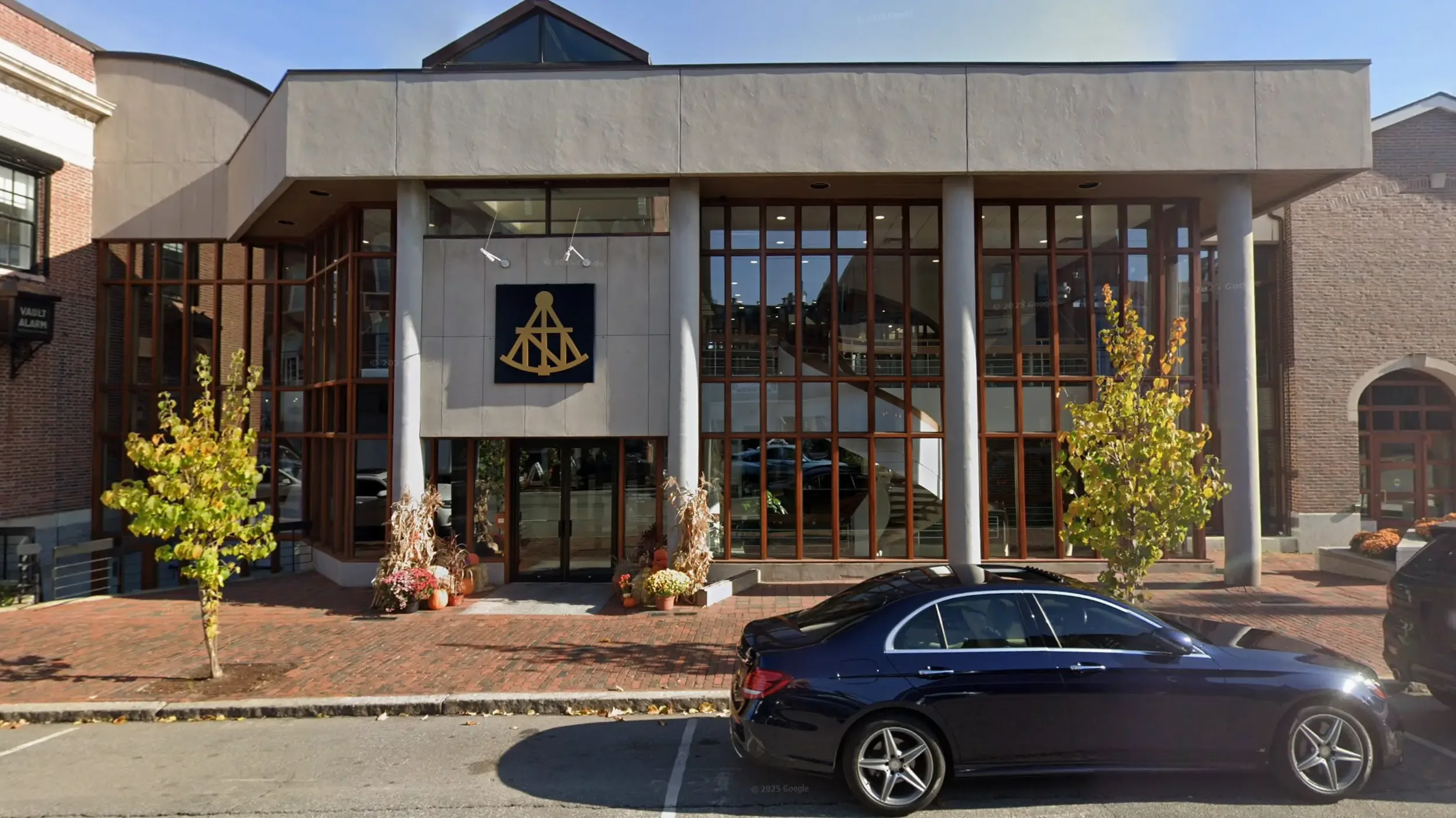 Modern two-story building with large glass windows, a nautical symbol above the entrance, fall decorations, and a parked black car in front on a brick-paved sidewalk.