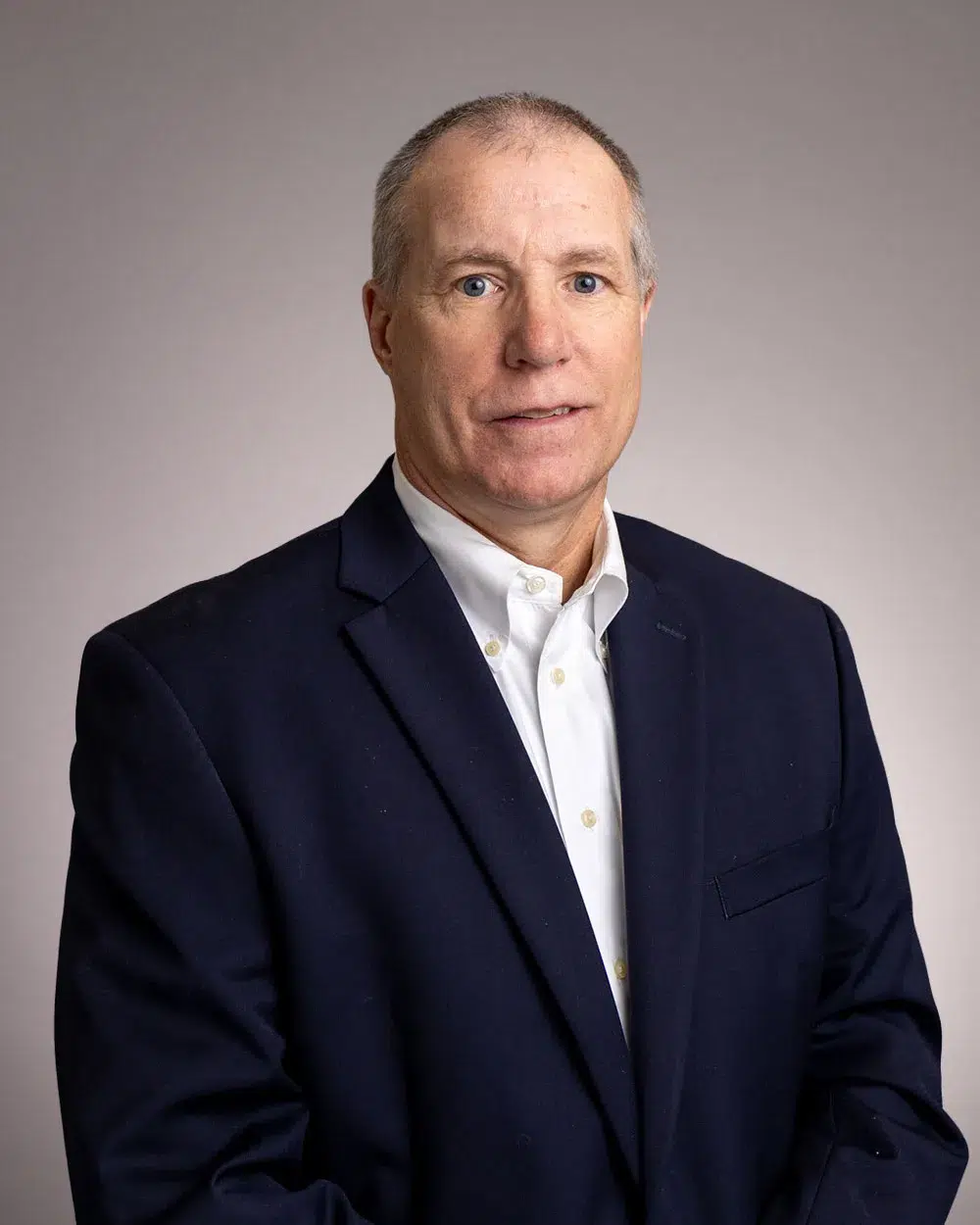 A man in a dark suit jacket and white dress shirt poses against a plain, light background, looking directly at the camera.