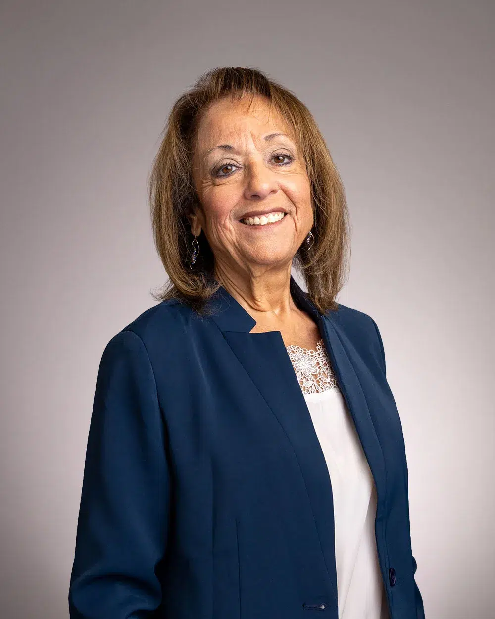 A woman with shoulder-length brown hair wearing a navy blue blazer and a white top, smiling and standing against a plain, light gray background.