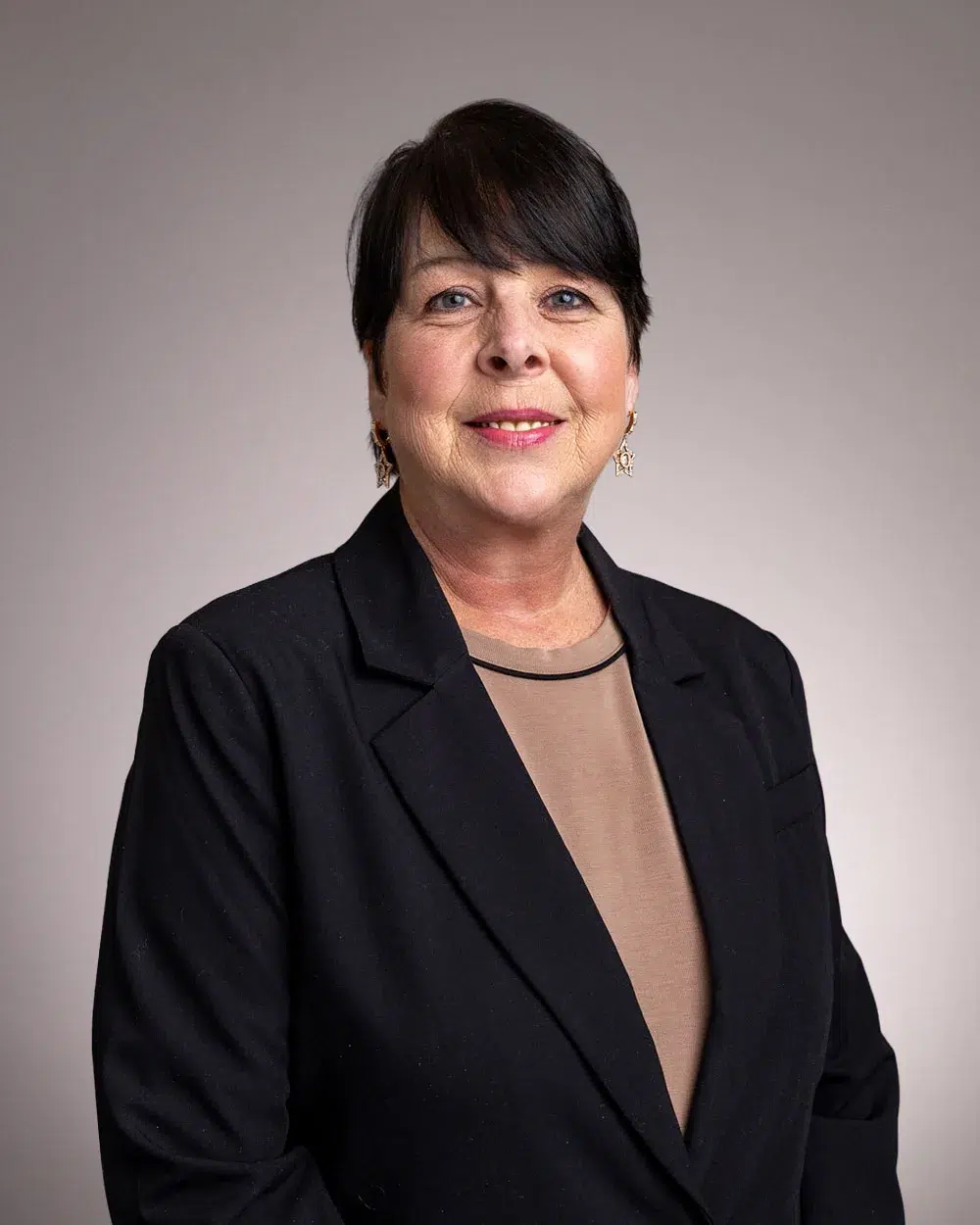 A woman with short dark hair, wearing a black blazer over a beige top, smiles at the camera against a plain light-colored background.