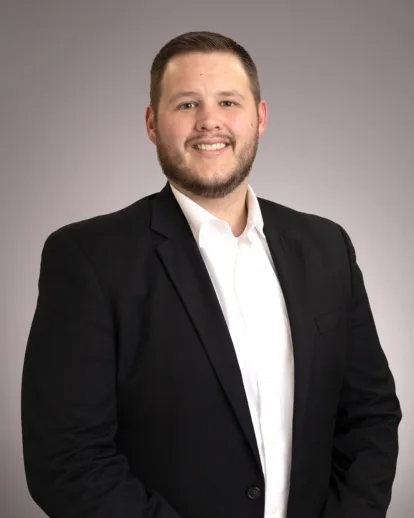 A man wearing a black suit jacket and white shirt stands smiling against a plain, light gray background.