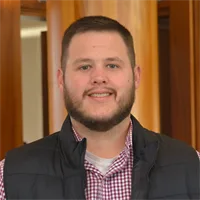 A man with short brown hair and a beard, wearing a black vest over a red and white checked shirt, smiles while standing indoors with wooden panels in the background.